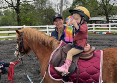 A young child wearing a riding helmet and pink boots sits on a brown pony with a maroon saddle pad, proudly holding a colorful ribbon. An adult stands beside them, smiling, with trees and a white fence in the background.