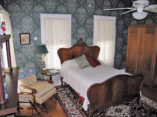guestroom A vintage bedroom with a wooden bed, white bedspread, patterned wallpaper, two windows with lace curtains, an armchair, a wooden wardrobe, a lamp, and a decorative rug on a wooden floor.