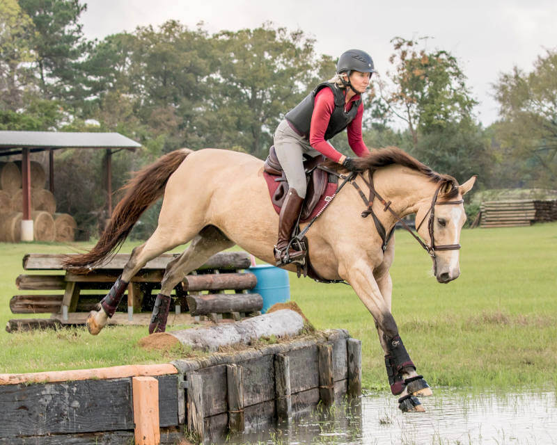 A person wearing a helmet and riding gear jumps a tan horse over a wooden obstacle above water on a grassy field during an equestrian event. Trees and hay bales are visible in the background.
