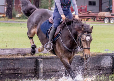 A person wearing a helmet and protective vest rides a dark horse as it jumps into a shallow water obstacle, splashing water, with grass, trees, and a barn in the background.