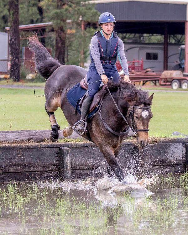A person wearing a helmet and protective vest rides a dark horse as it jumps into a shallow water obstacle, splashing water, with grass, trees, and a barn in the background.