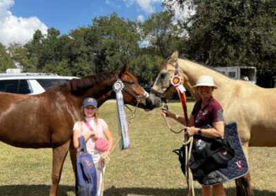 Two people stand outdoors holding horses with award ribbons. A girl in a pink cap and a woman in a wide-brim hat smile, each holding a horse. Trees and blue sky are visible in the background.