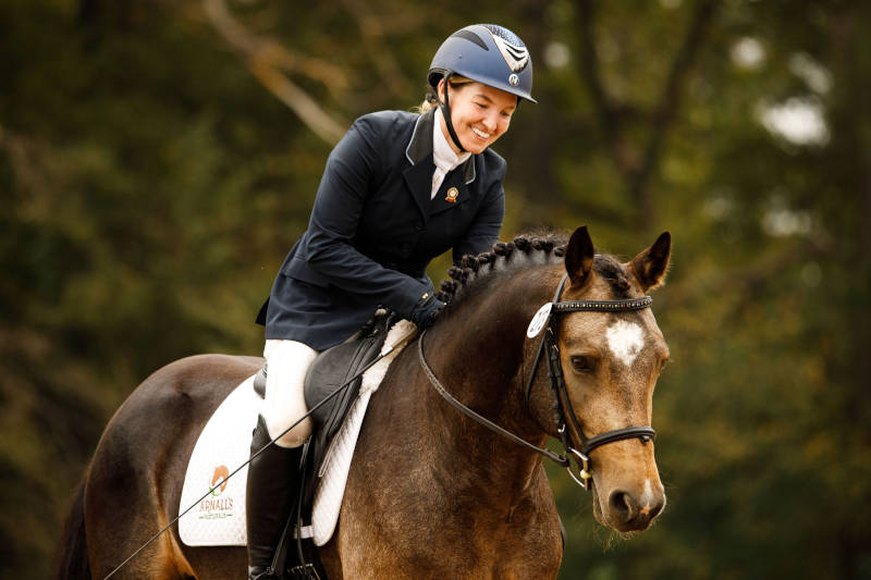 A smiling equestrian in a navy helmet and riding jacket gently pats the neck of a brown horse with a white patch on its forehead during an outdoor event. Trees are visible in the blurred background.